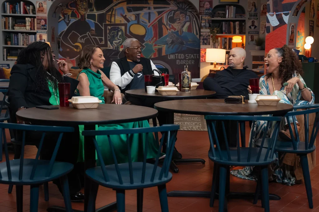 Returning A Different World characters talk around tables beneath a large mural