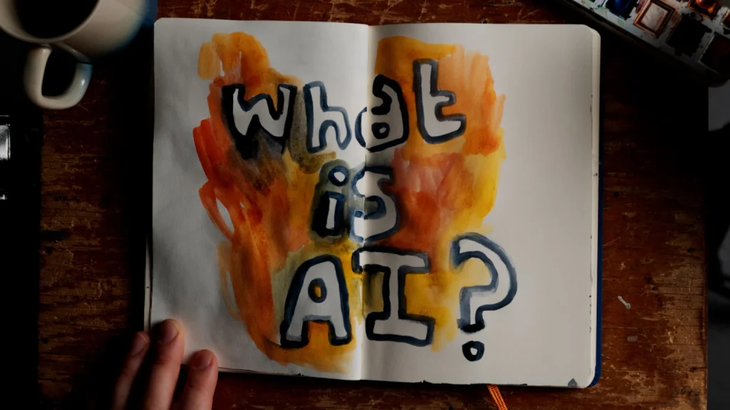 An open watercolor notebook reading What is AI? on a wooden desk beside a mug and paint palette