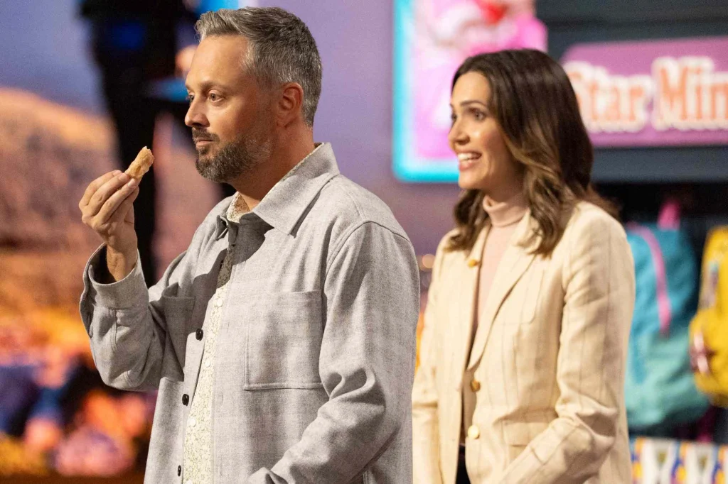 Nate Bargatze holds a snack beside Mandy Moore on the Shark Tank set in The Breadwinner