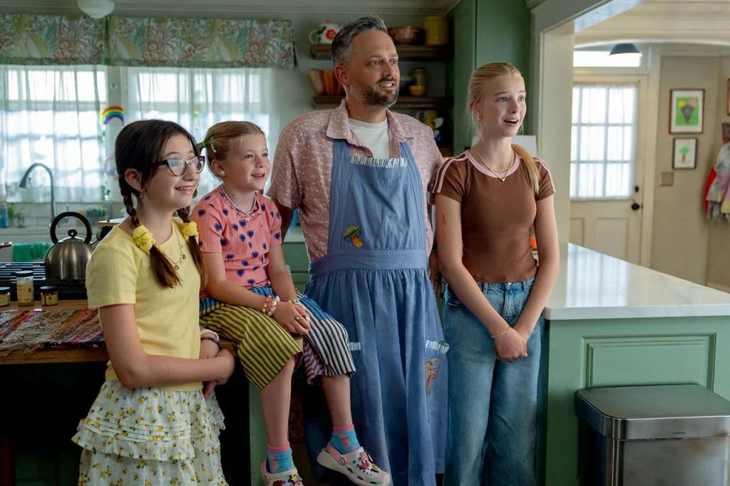 Nate Bargatze as Nate Wilcox stands in a blue apron with his three daughters in a kitchen scene from The Breadwinner