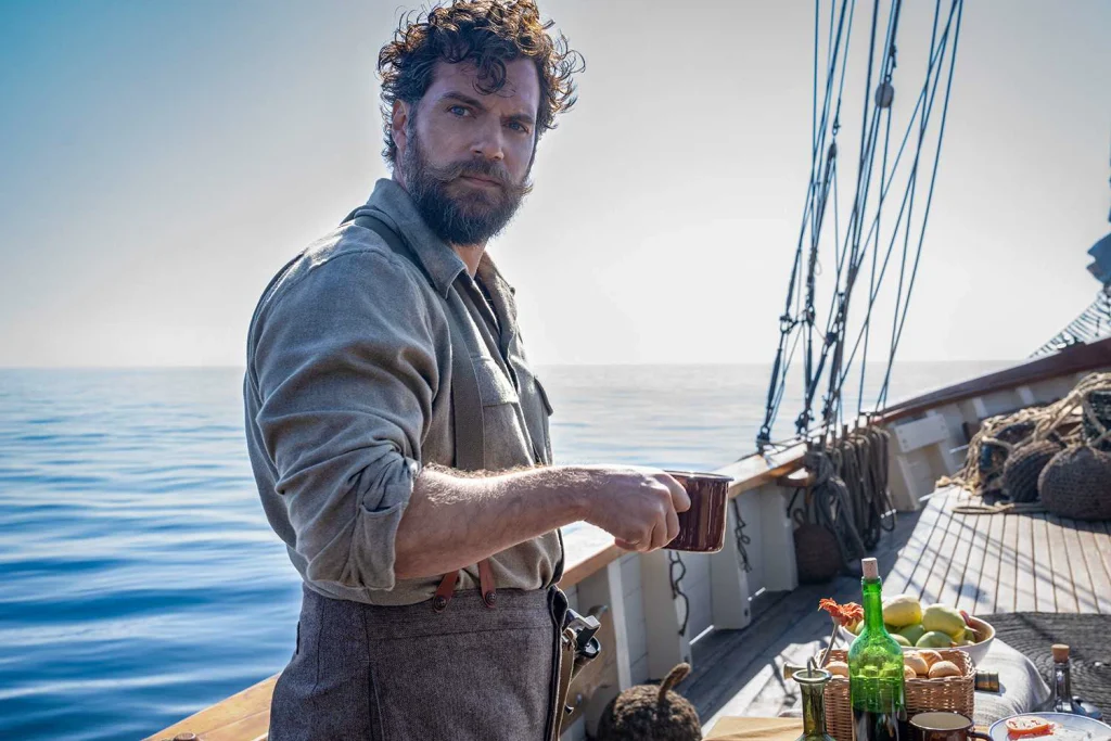 A bearded man holding a mug on a wooden boat, with breakfast dishes and open water behind him
