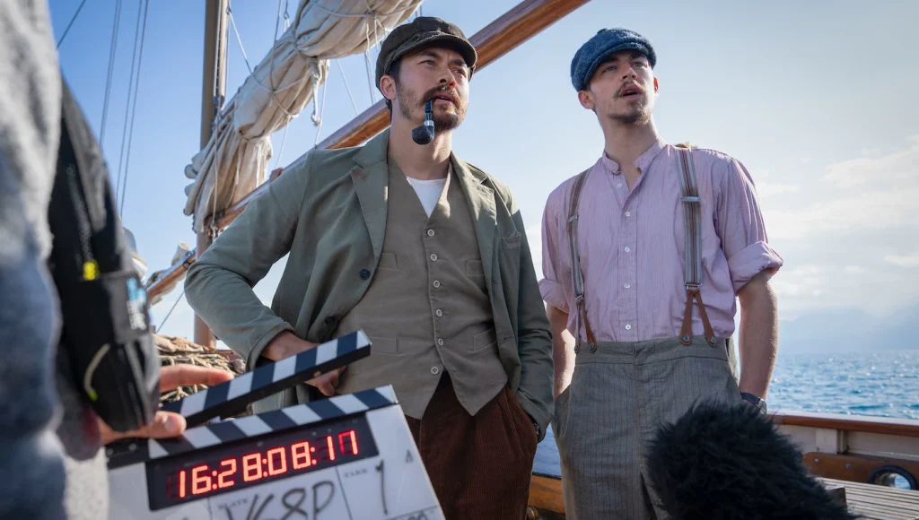Henry Golding and Hero Fiennes Tiffin on a boat, with a clapperboard and microphone visible in the foreground