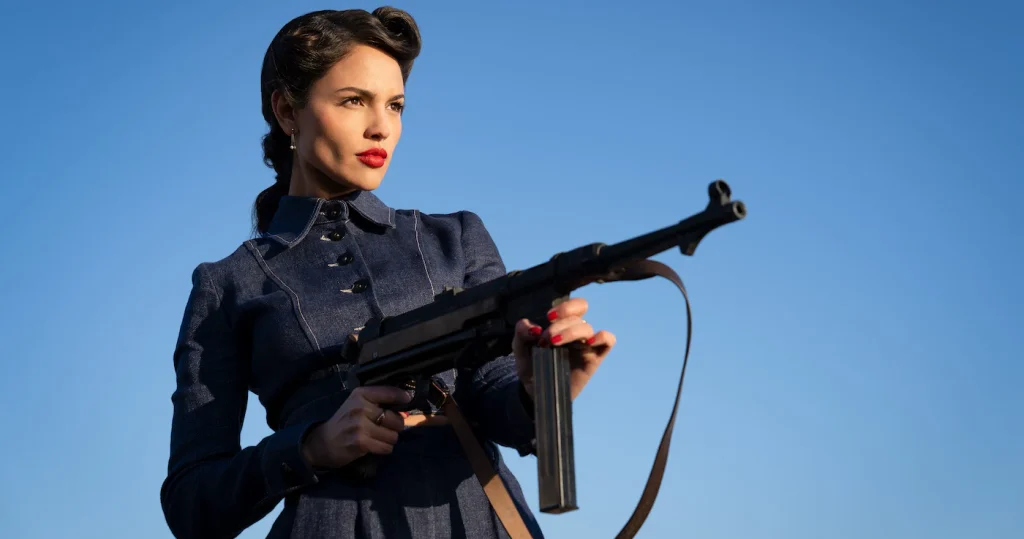 Eiza González holding a submachine gun against a blue sky, with warm light across her face