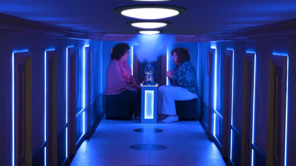 Two contestants sit opposite each other beside a gumball machine in a blue-lit corridor in Wonka's The Golden Ticket