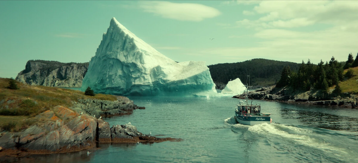 A fishing boat approaches a large iceberg between rocky, wooded shores in Below.