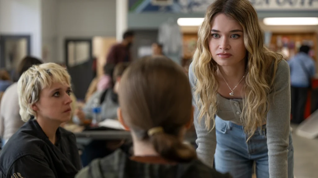 Emaline sits beside Carrie as Chris leans toward their cafeteria table.