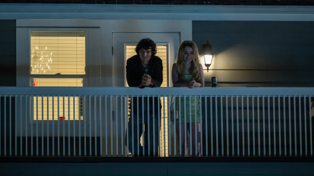 Billy and Chris lean on a porch railing outside a lit house at night.
