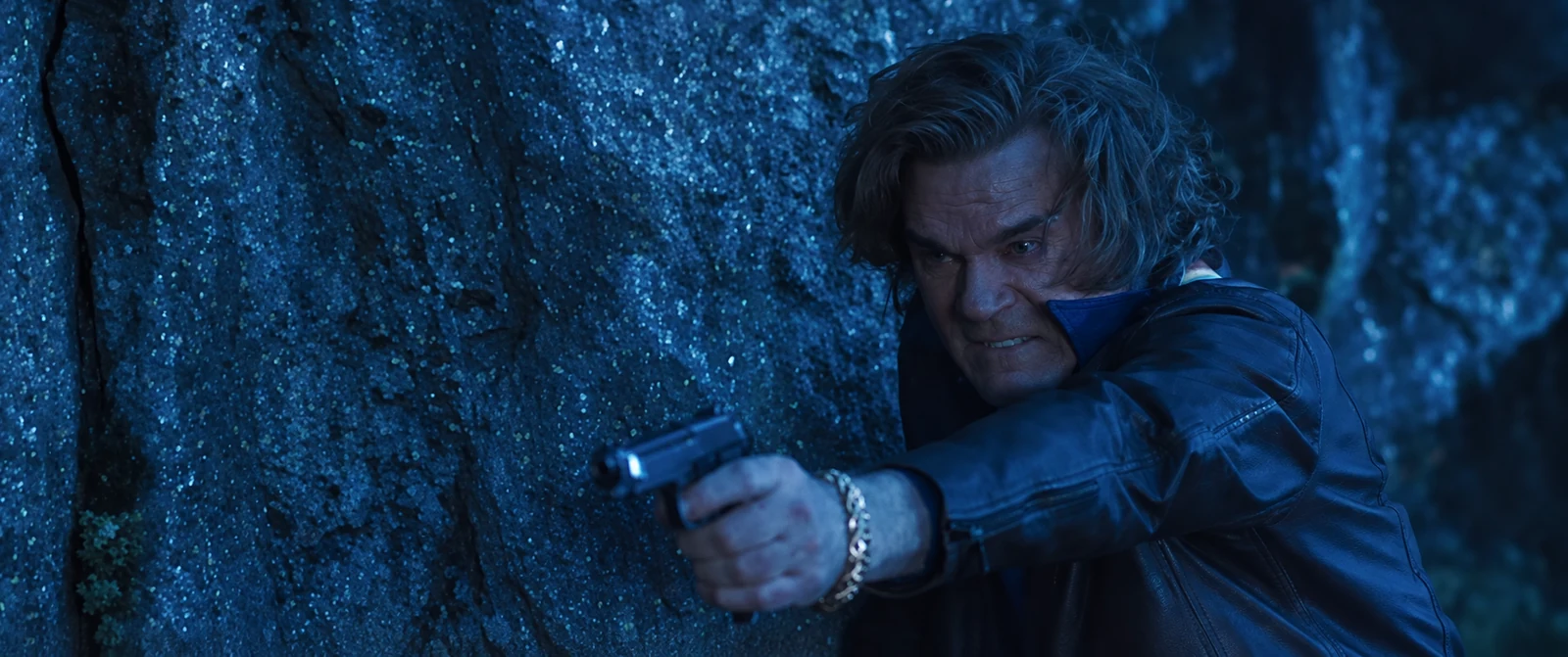 A man in a leather jacket aims a handgun beside a tree in Cocaine Bear