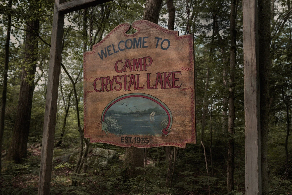 A wooden Camp Crystal Lake welcome sign hangs among trees.
