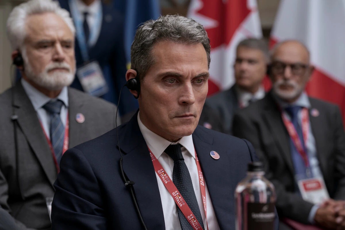 Hal Wyler wears an earpiece and a G7 badge while seated among delegates in The Diplomat