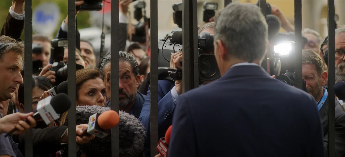 Hal Wyler faces reporters with microphones and cameras on the other side of an iron gate