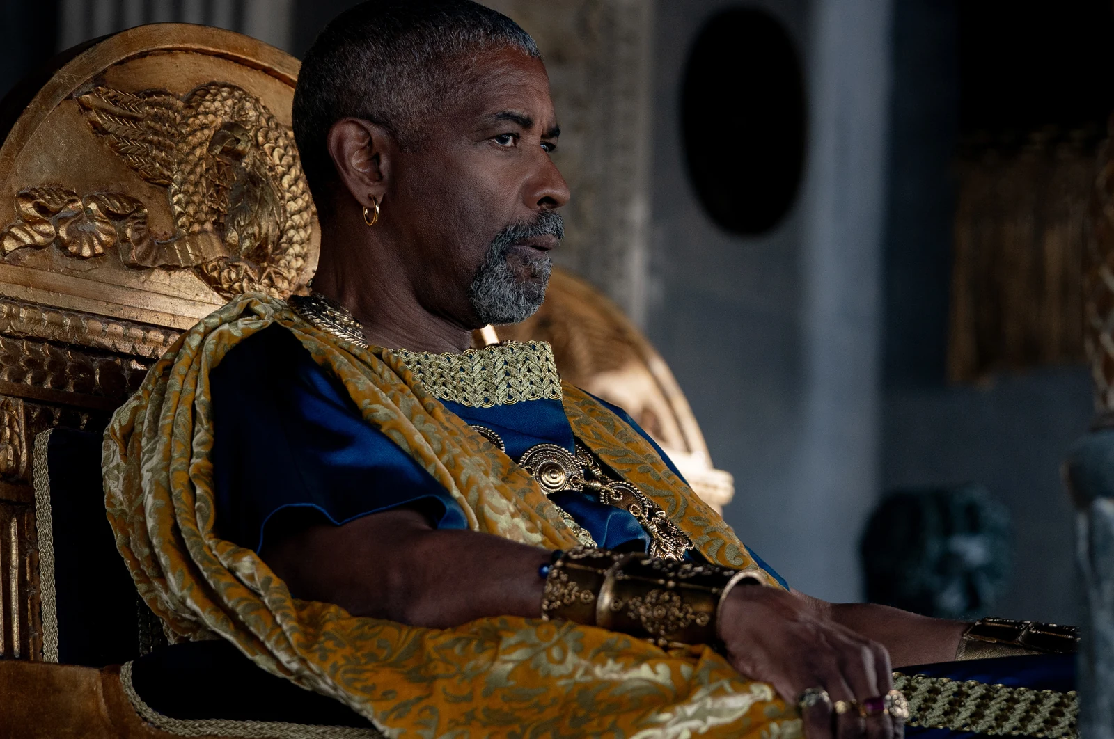 Macrinus sits in an ornate chair wearing blue and gold robes in Gladiator II.