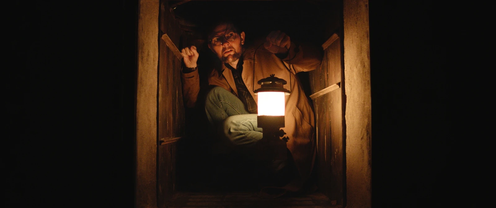 Ohm Bauman crouches in a narrow wooden opening with a lit lantern in Hokum.