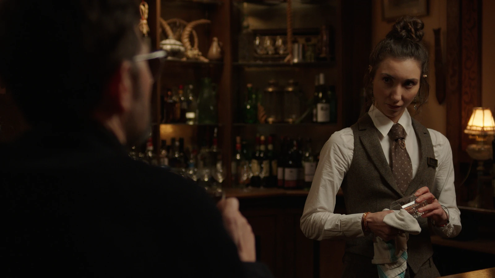 A hotel bartender polishes a drinking glass while facing Ohm in Hokum.