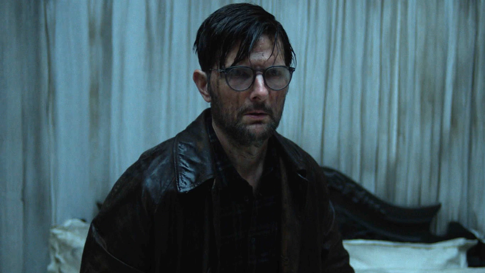 Ohm Bauman wears glasses and a dark jacket in front of pale curtains in Hokum.