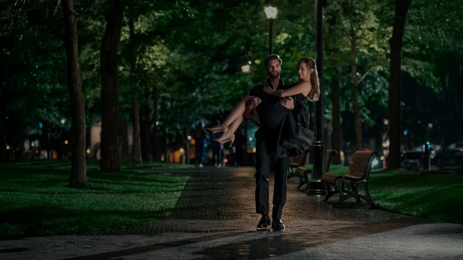 Adam carries Olive along a tree-lined path at night.