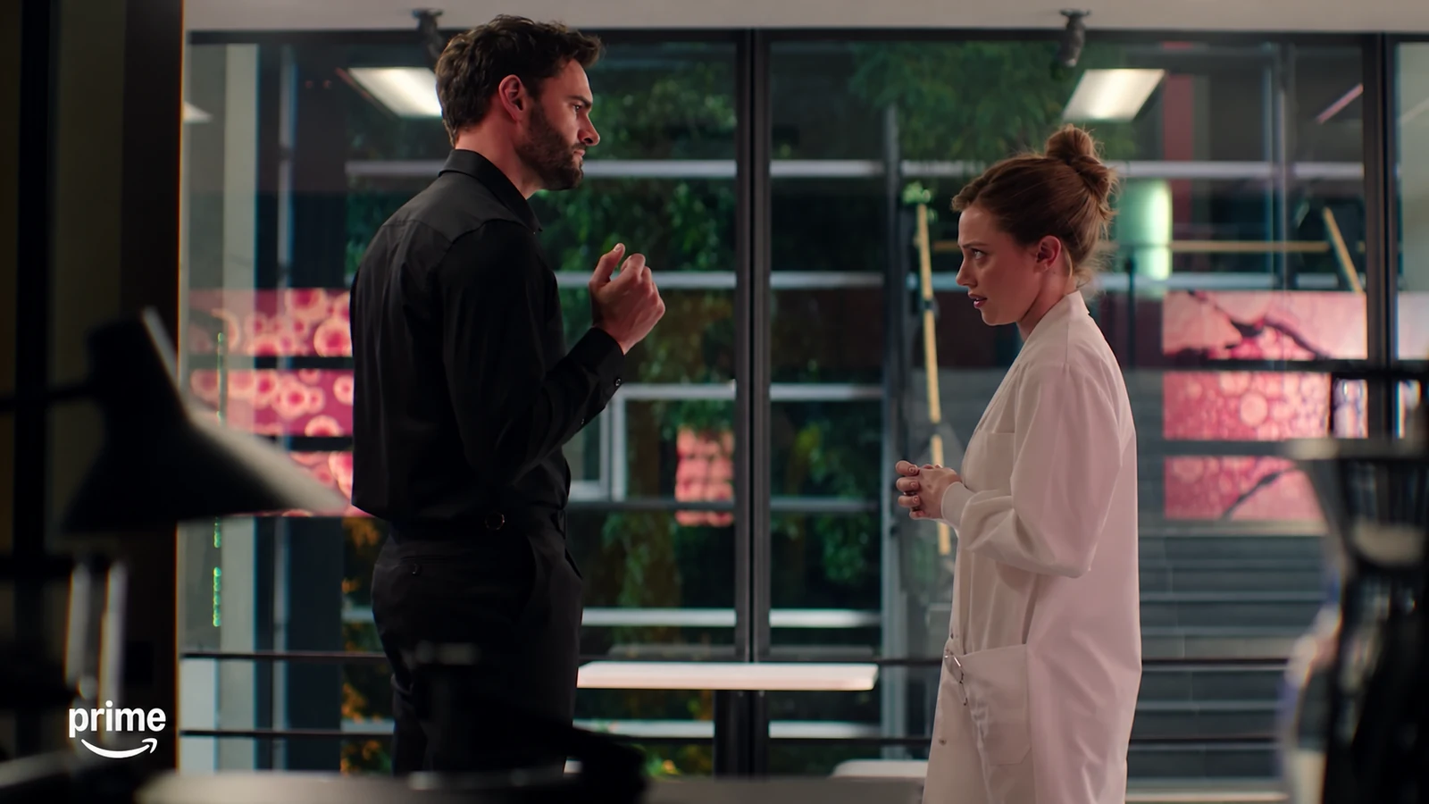 Olive, wearing a white lab coat, faces Adam beside a wall of windows.
