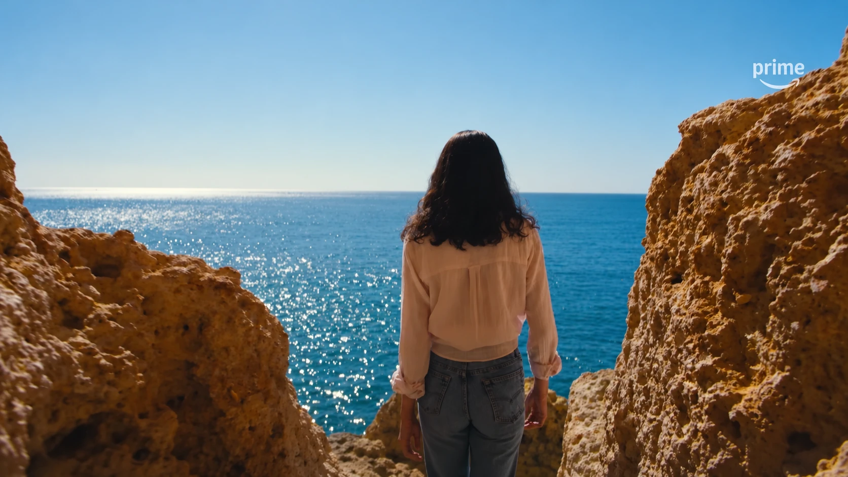 A woman seen from behind looks toward the sea between sunlit rocks