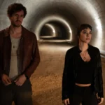 Victor Belmondo and Simona Tabasco stand side by side in a lit tunnel in Masterplan.