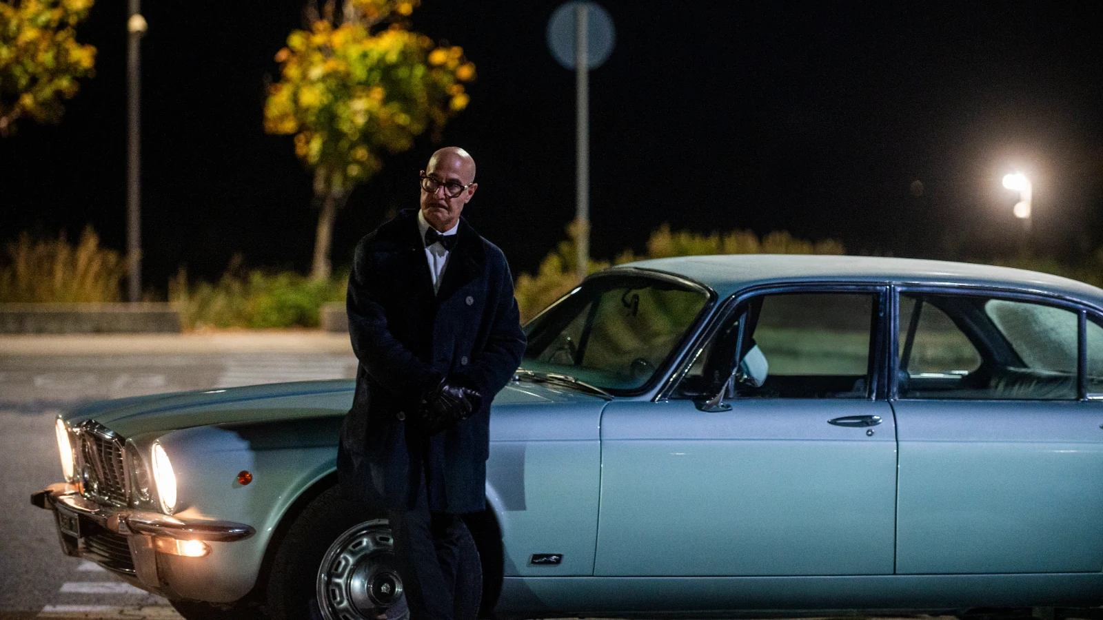 Stanley Tucci stands beside a pale blue car at night, wearing a dark overcoat and gloves.