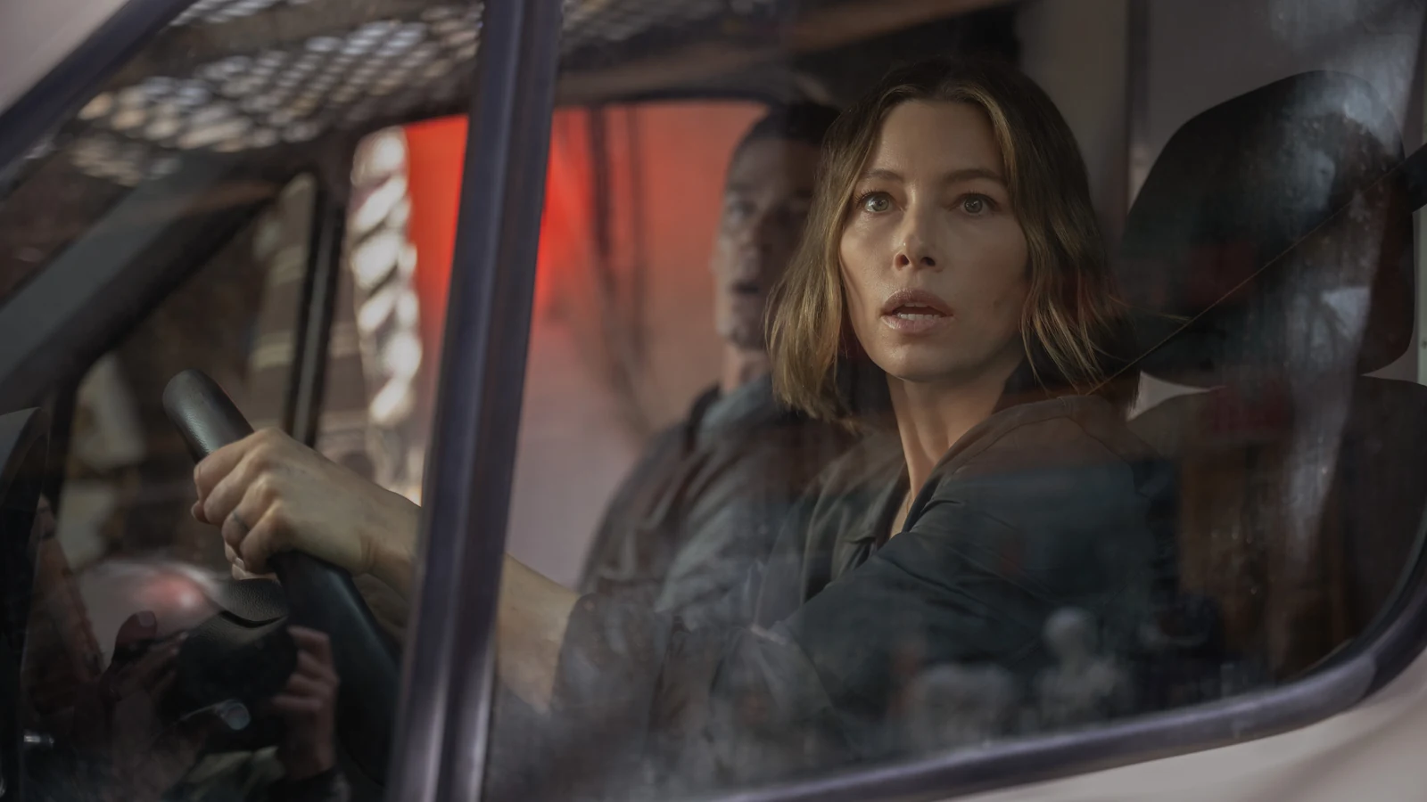 Aimee holds the steering wheel with Sean beside her, seen through a vehicle window.