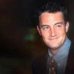 Matthew Perry smiles in a dark jacket, blue shirt, and patterned tie.