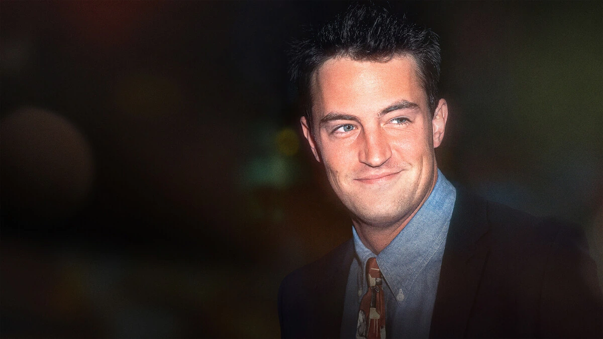 Matthew Perry smiles in a dark jacket, blue shirt, and patterned tie.