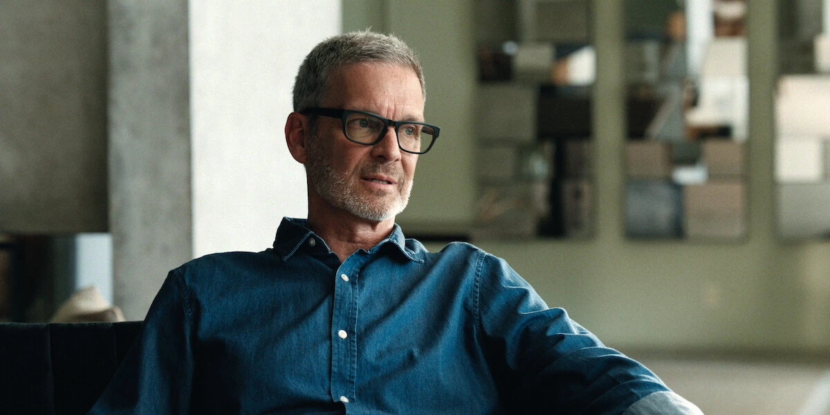 A man wearing black glasses and a blue shirt sits on a sofa in The One About Matthew Perry.