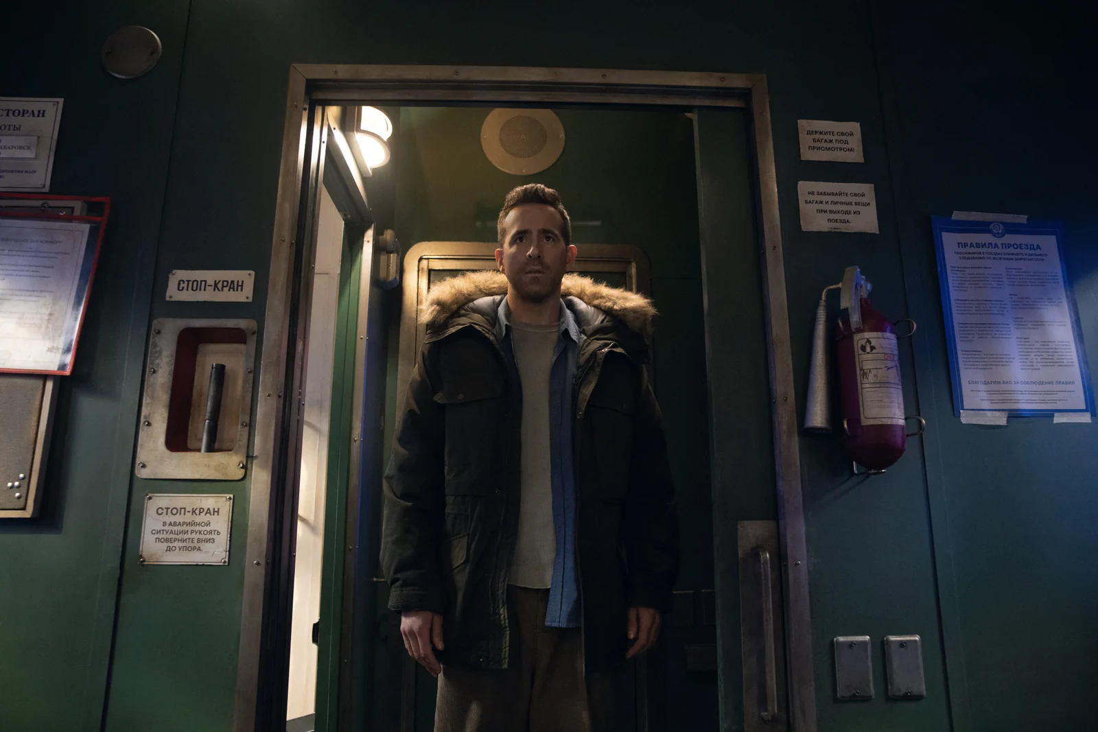 Troy stands in a train doorway wearing a fur-trimmed winter coat.