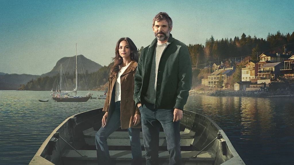 Two people stand in a small boat with a harbor and hillside town behind them in Murder in a Small Town