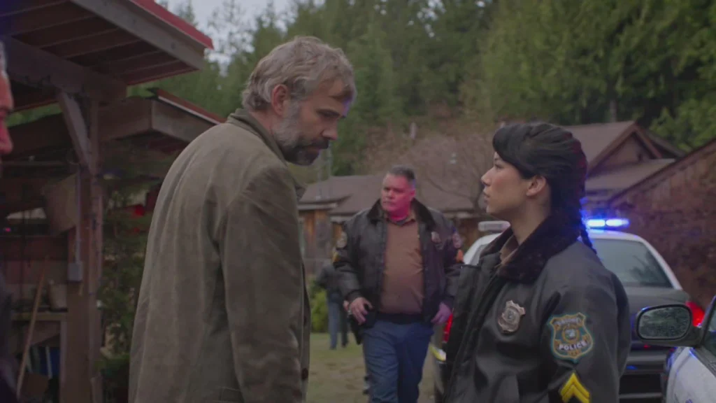 A man faces a uniformed officer beside police vehicles and wooden buildings in Murder in a Small Town