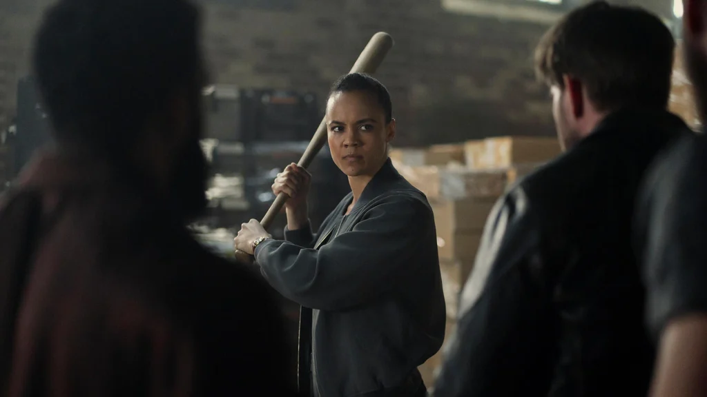 Frances Neagley holds a baseball bat over her shoulder, facing two figures in the foreground.