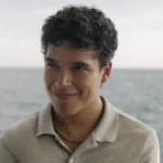 A young man in a gray polo shirt smiles with the sea behind him in Surviving Hurricane Otis.