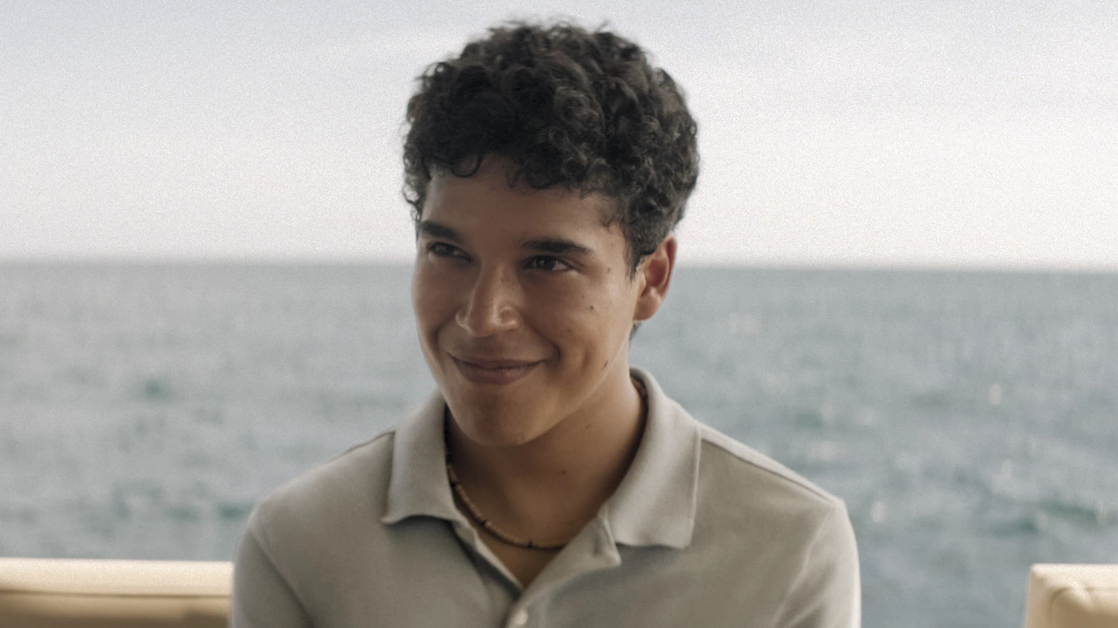 A young man in a gray polo shirt smiles with the sea behind him in Surviving Hurricane Otis.