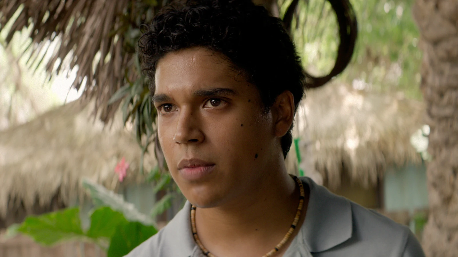 A young man with curly hair wears a gray polo shirt beneath a thatched roof in Surviving Hurricane Otis.