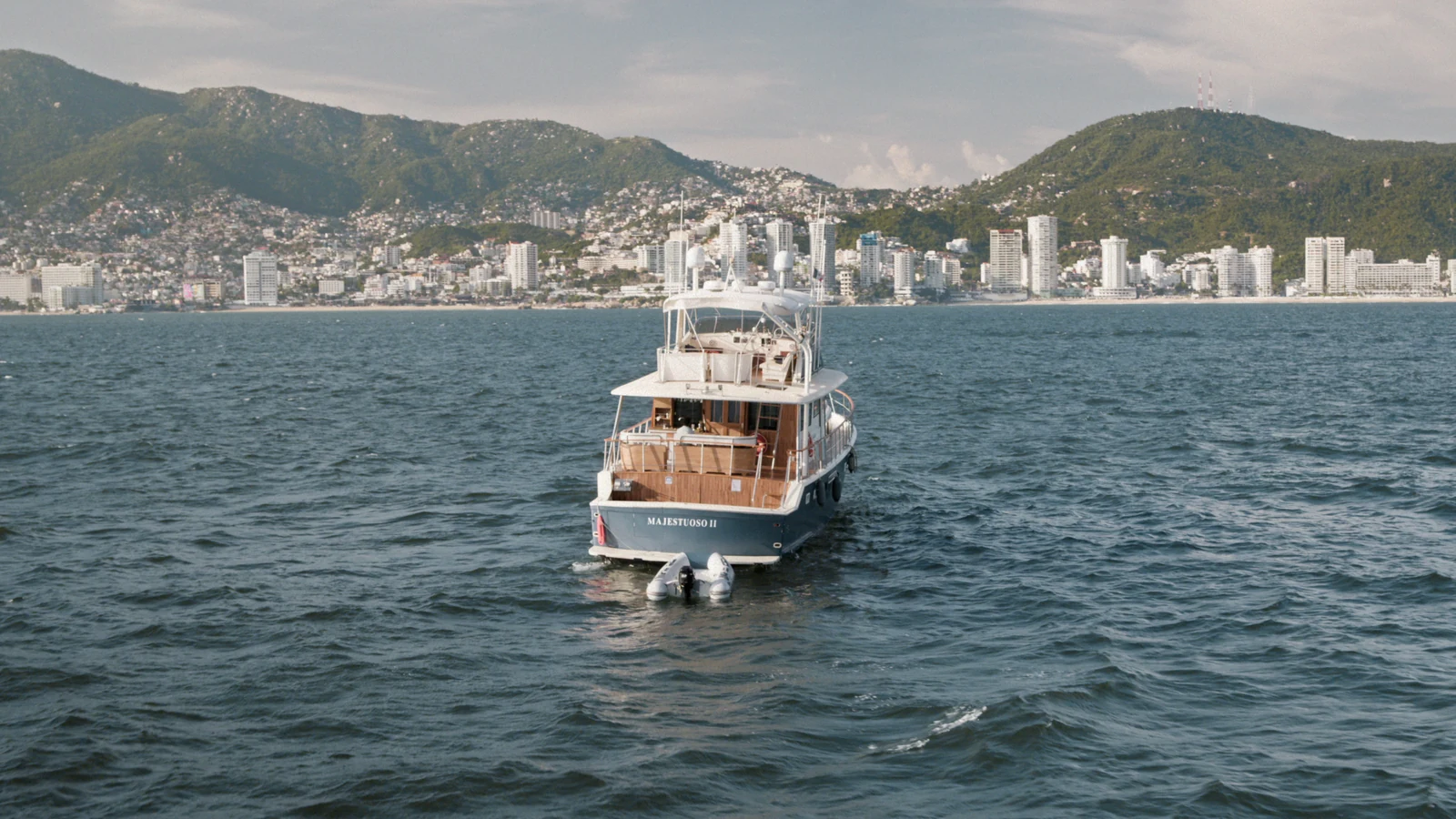 A yacht crosses the water with a coastal city and green hills behind it in Surviving Hurricane Otis.