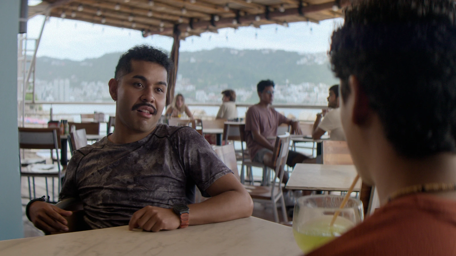 Two men sit across a table at an open-air waterfront restaurant in Surviving Hurricane Otis.