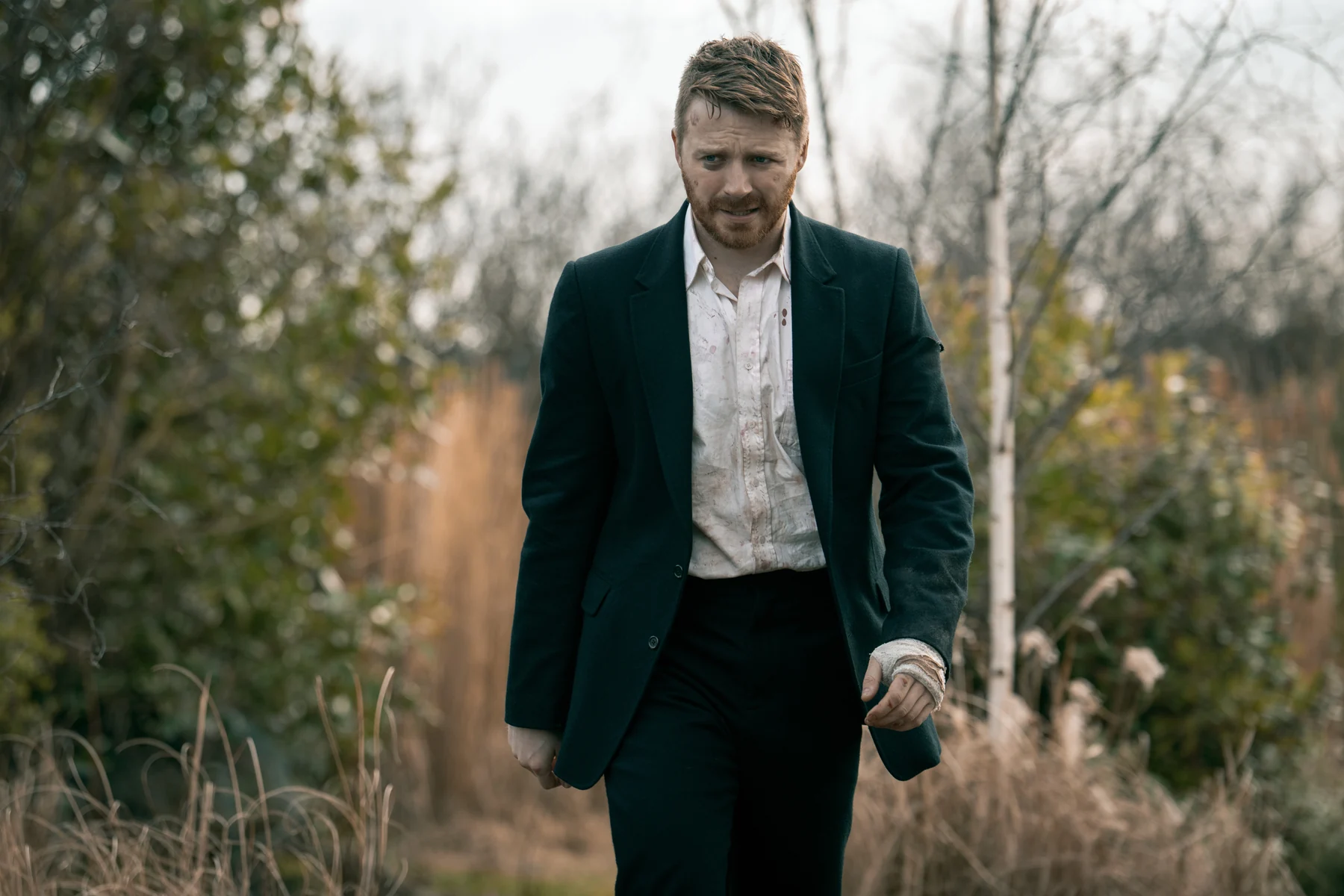 Jack Lowden as River Cartwright walks outdoors in a dark suit with a bandaged hand.