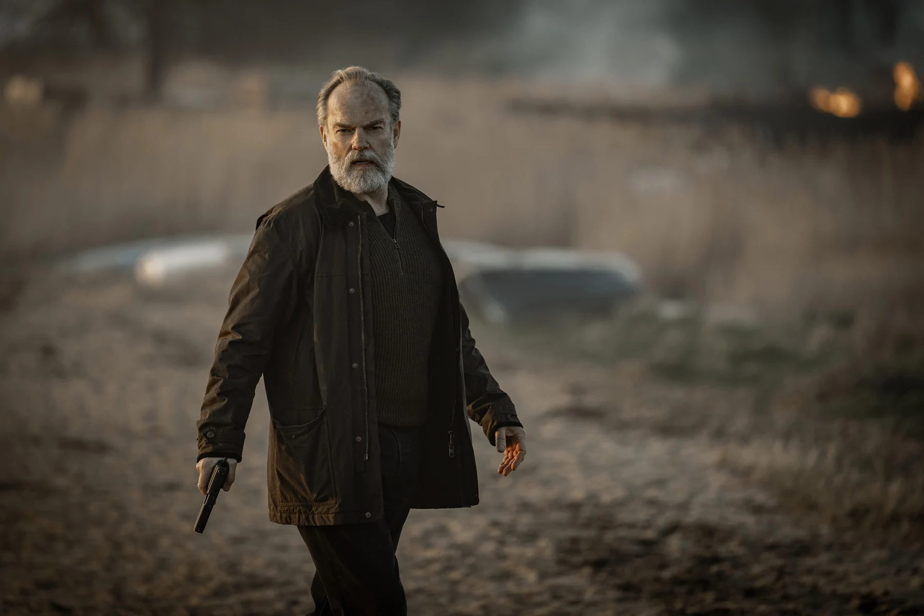 Hugo Weaving as Frank Harkness holds a handgun beside a field of dry grass.