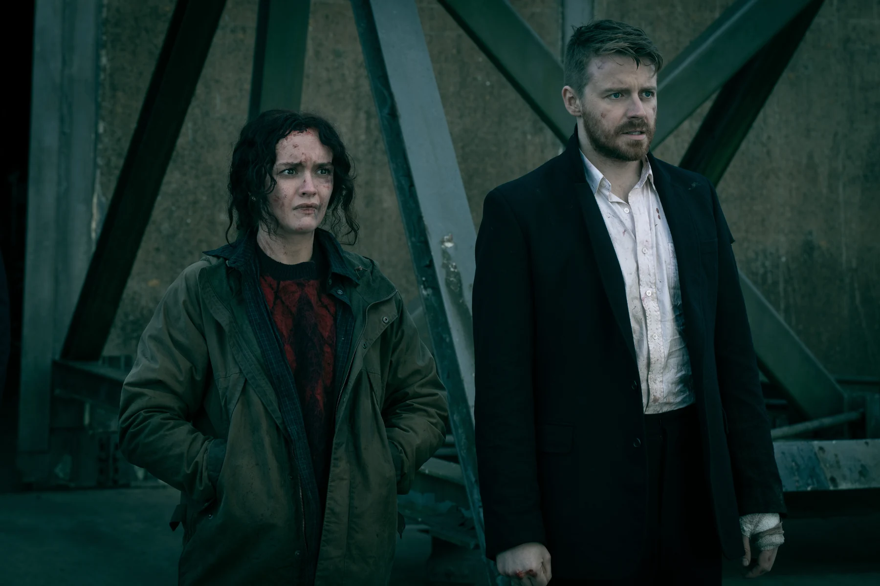 Olivia Cooke as Sid Baker stands beside Jack Lowden as River Cartwright against a metal structure.
