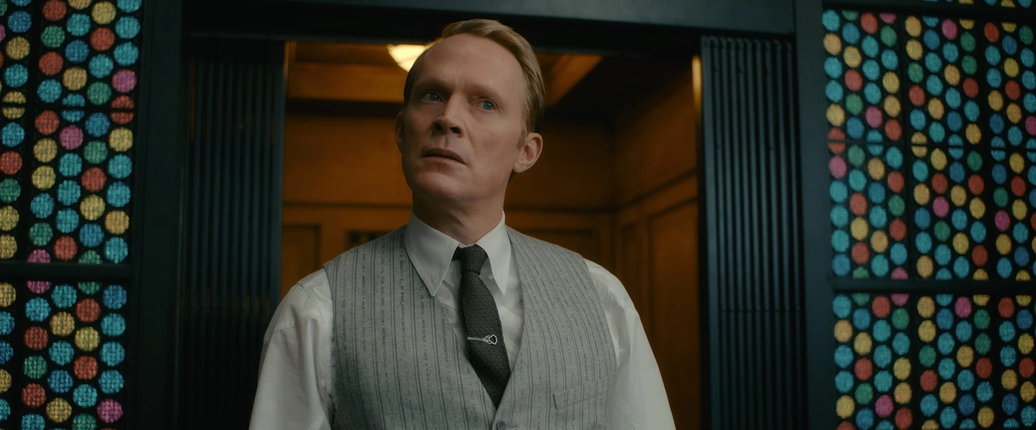 Paul Bettany as Vision in a gray vest beside colored glass panels