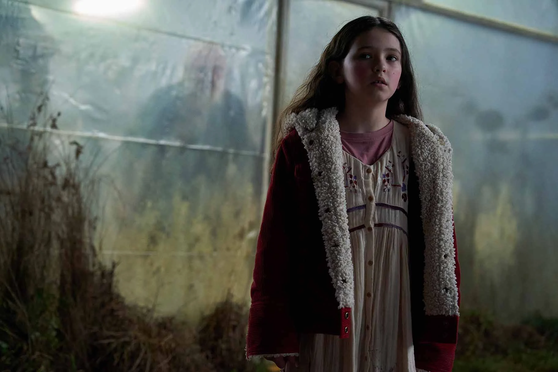 Ginger stands in front of a translucent wall, wearing a red jacket with a cream lining in Wolf Man.